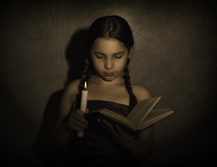little girl reading book and studying in candle light in dark room