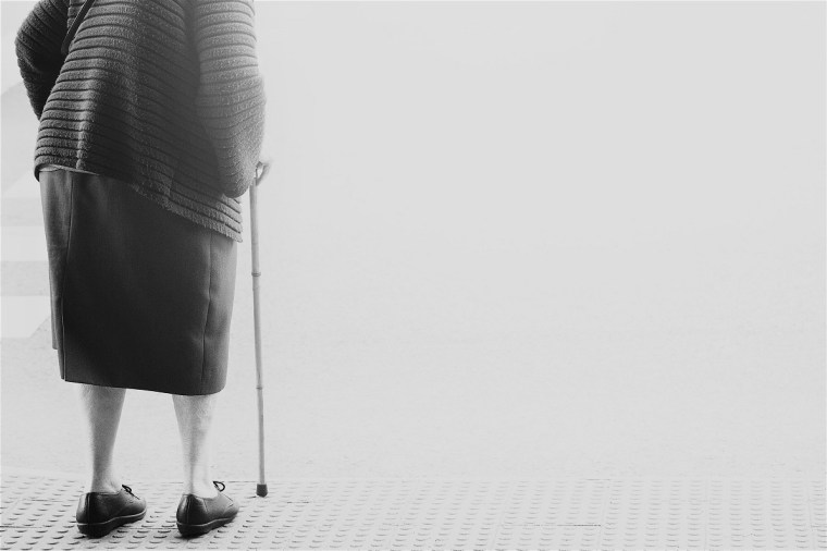 elderly old woman with walking stick stand waiting on footpath sidewalk crossing the street alone. concept senior across the street to zebra crosswalk.